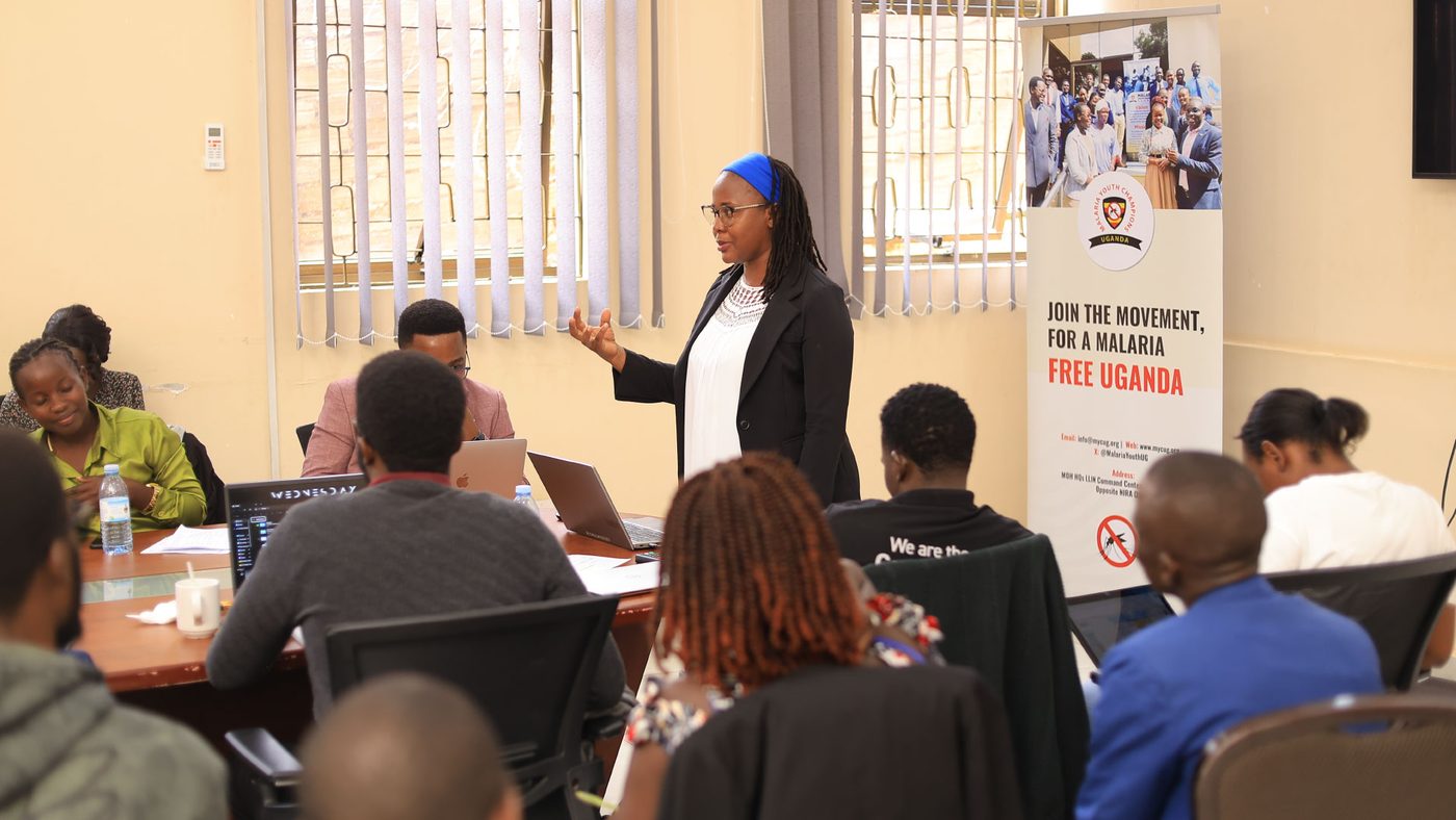 A facilitator speaks to young people at laptops during an MYCU workshop, beside a 'Join the movement for a malaria free Uganda' banner