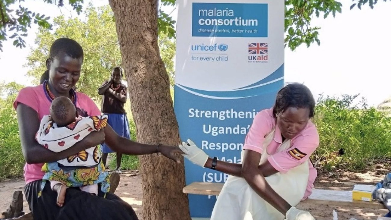 A health worker in gloves and an apron tests a mother for malaria as she holds her baby, beside a Strengthening Uganda’s Response to Malaria banner
