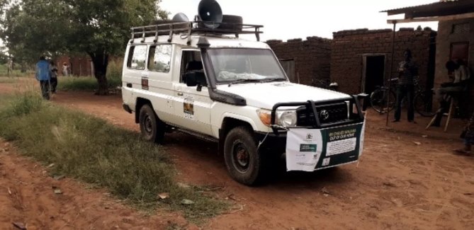 A white Land Cruiser fitted with loudspeakers for community health messaging parked in a village