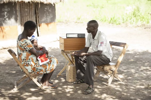 Village Health Team member Joseph Ewaru sits with a mother and her child beside his VHT kit outside a home in Ogerai Village