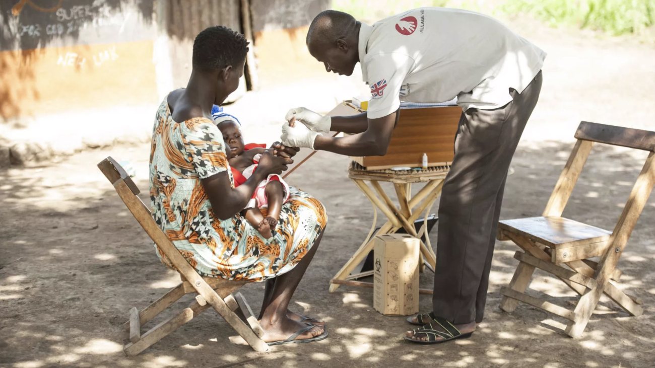 A Village Health Team member in gloves tests a baby for malaria as the mother holds the child on a chair outside their home