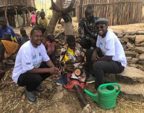 Two MYCU youth champions crouch beside a mother and child in a Karamoja homestead as the child is given SPAQ