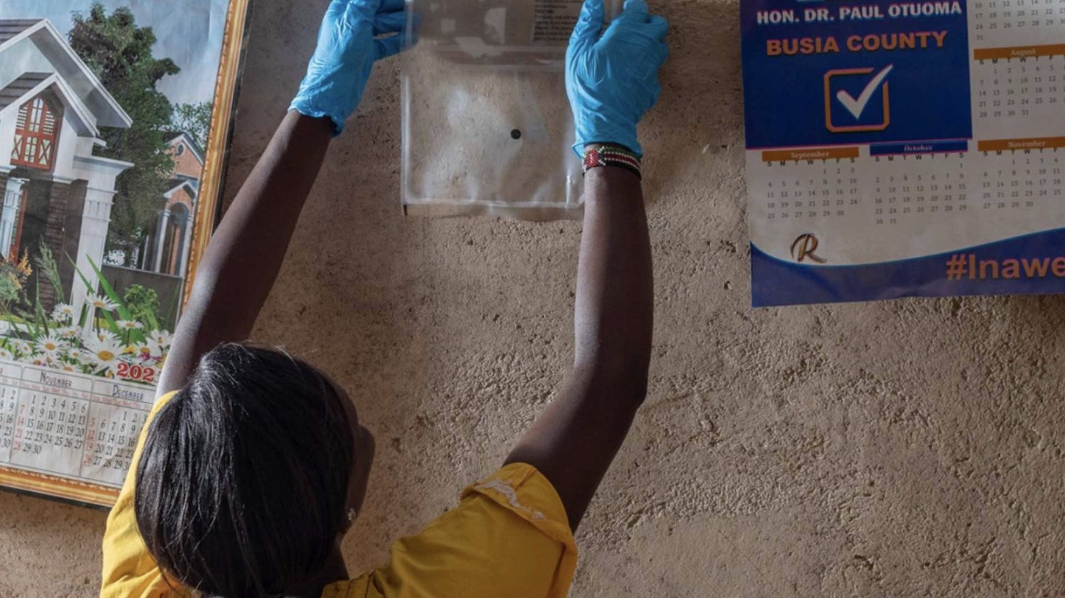 A community health worker in blue gloves fixes a spatial repellent to the wall of a home
