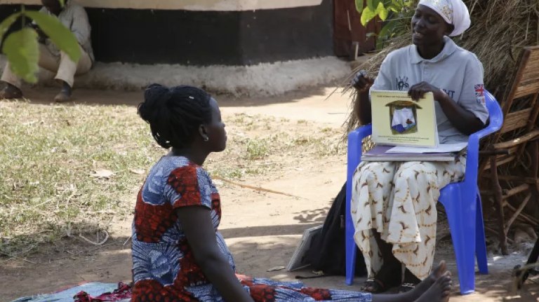 A Village Health Team member seated on a blue chair shows an illustrated malaria flipchart to a pregnant woman sitting on a mat outside a home