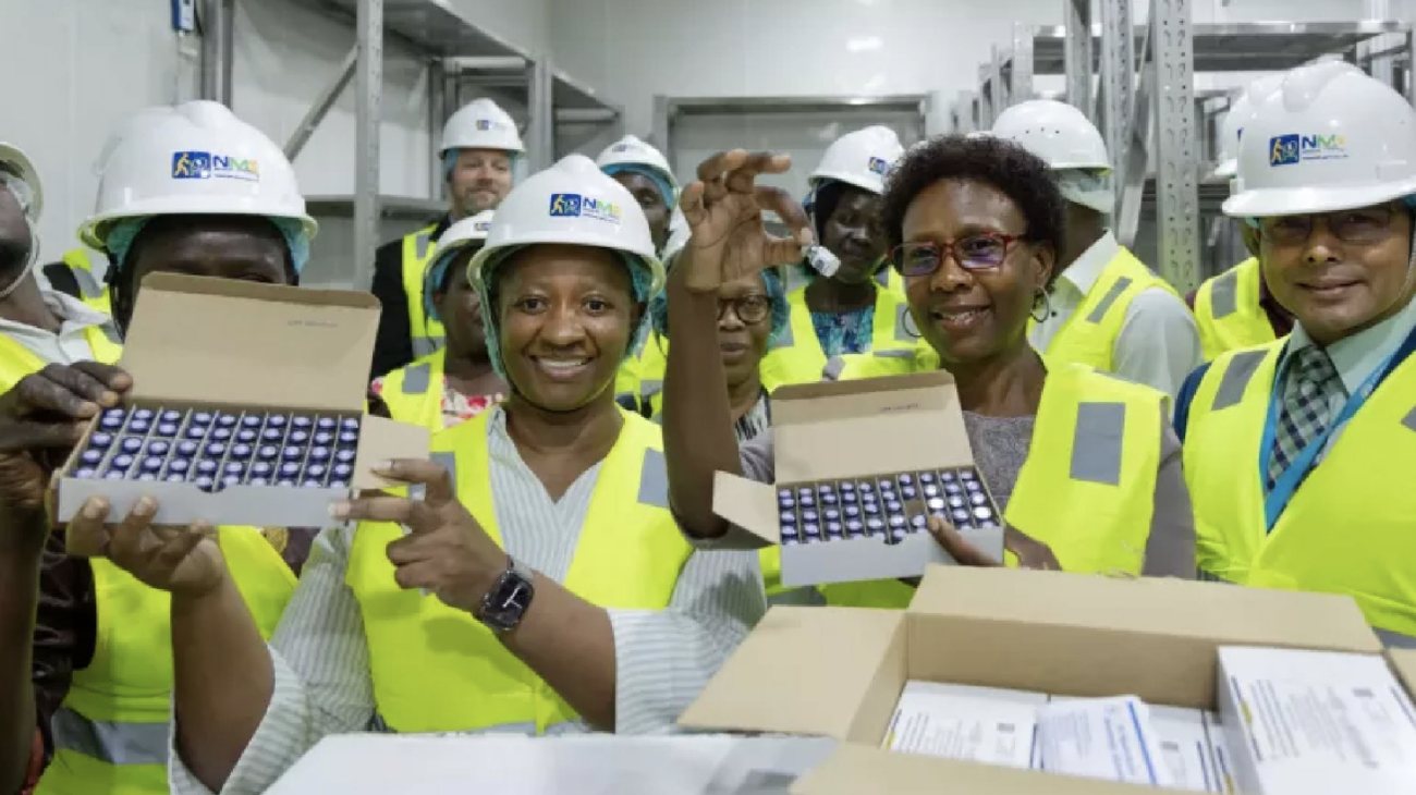 Health officials in hard hats and high-visibility vests smile and hold up trays of malaria vaccine vials at the National Medical Stores