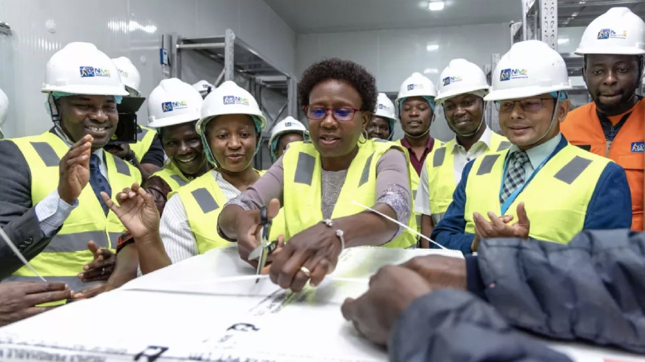 Health Minister Dr Jane Ruth Aceng, in a hard hat and high-visibility vest, cuts the seal on the first box of malaria vaccines at the National Medical Stores, surrounded by smiling officials