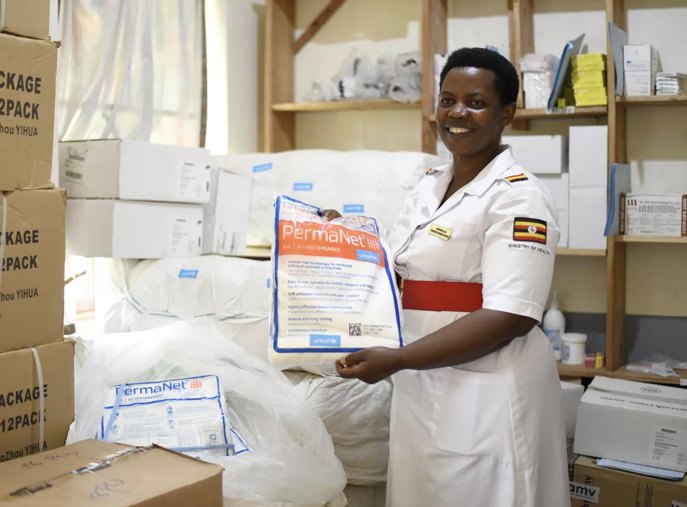 A health worker in a white uniform holds up a PermaNet mosquito net in a health centre store stacked with boxes of nets