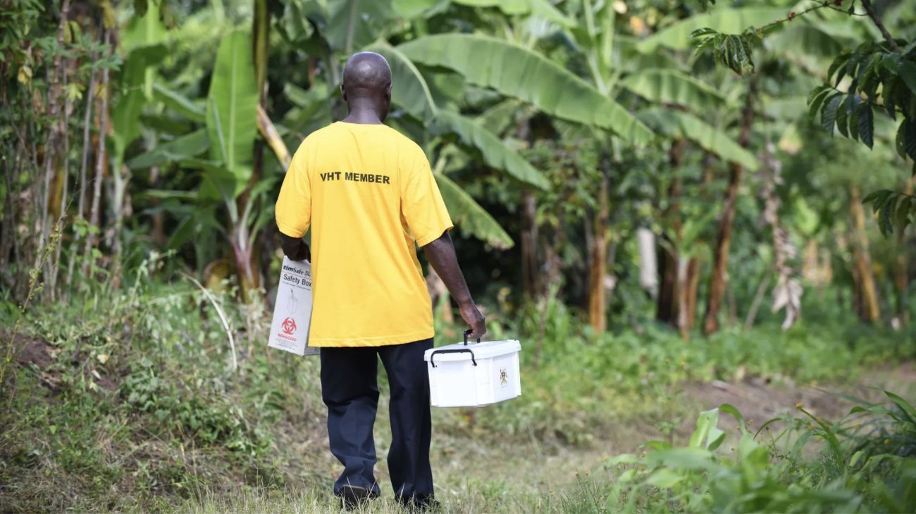 A Village Health Team member in a yellow VHT Member T-shirt walks through banana plantations carrying a medicine box and a safety box