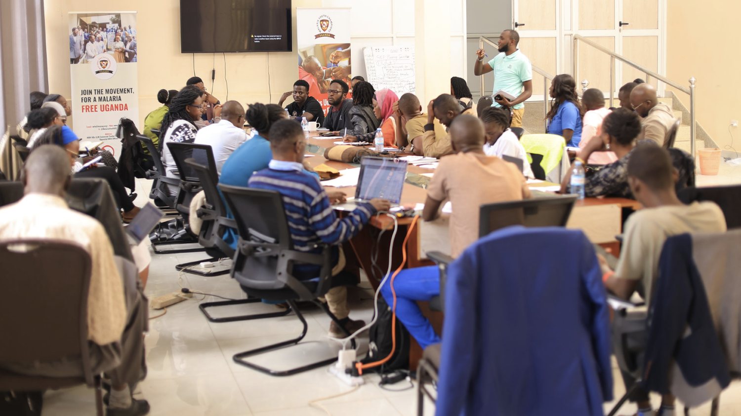 Young people work on laptops around a long conference table at an MYCU workshop, beside a Join the movement, for a malaria free Uganda banner