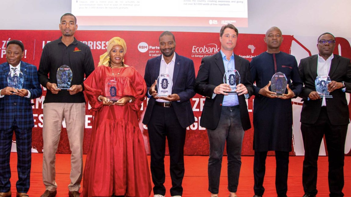 Seven award winners stand on stage holding glass trophies in front of a red Zero Malaria Business Leadership Initiative backdrop with Ecobank, RBM Partnership and Speak Up Africa logos